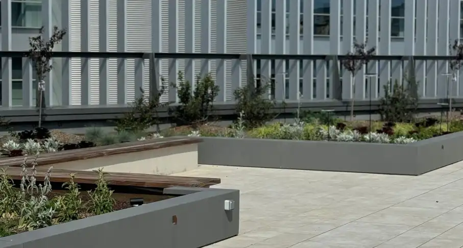 Terraza ajardinada con jardineras grises y pavimento claro junto a un edificio de oficinas